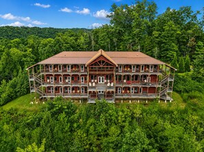A large, multi-story wooden building with balconies, surrounded by lush greenery and a forested backdrop. - Foxwood Mansion 14 Bedroom Cabin by RedAwning (Sevierville)