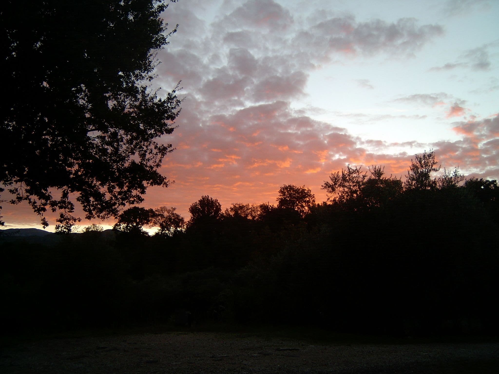 Un atardecer con nubes, árboles y el silueta de colinas al fondo.
