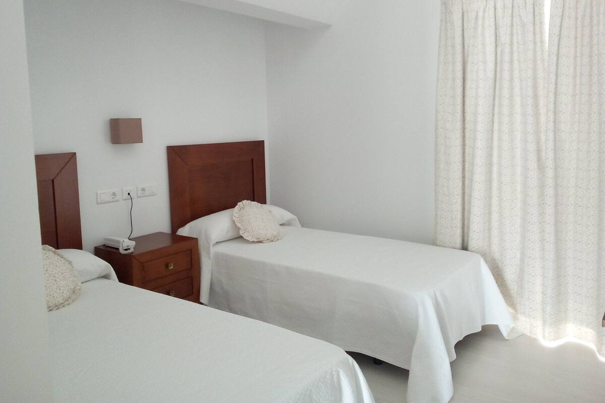 Une chambre d’hôtel avec deux lits simples, une table de chevet en bois, une lampe fixée au mur et une grande fenêtre avec des rideaux.