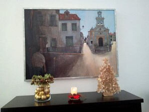 Una pintura de una escena callejera con edificios y una fuente, un jarrón con adornos dorados, una vela y un árbol decorativo sobre una mesa negra.