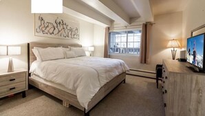 Un dormitorio con una cama grande, una mesita de noche, un aparador, un televisor y una ventana con cortinas.