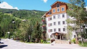Un edificio de varios pisos con fachada blanca y detalles de madera, rodeado de abundante vegetación y un paisaje montañoso.