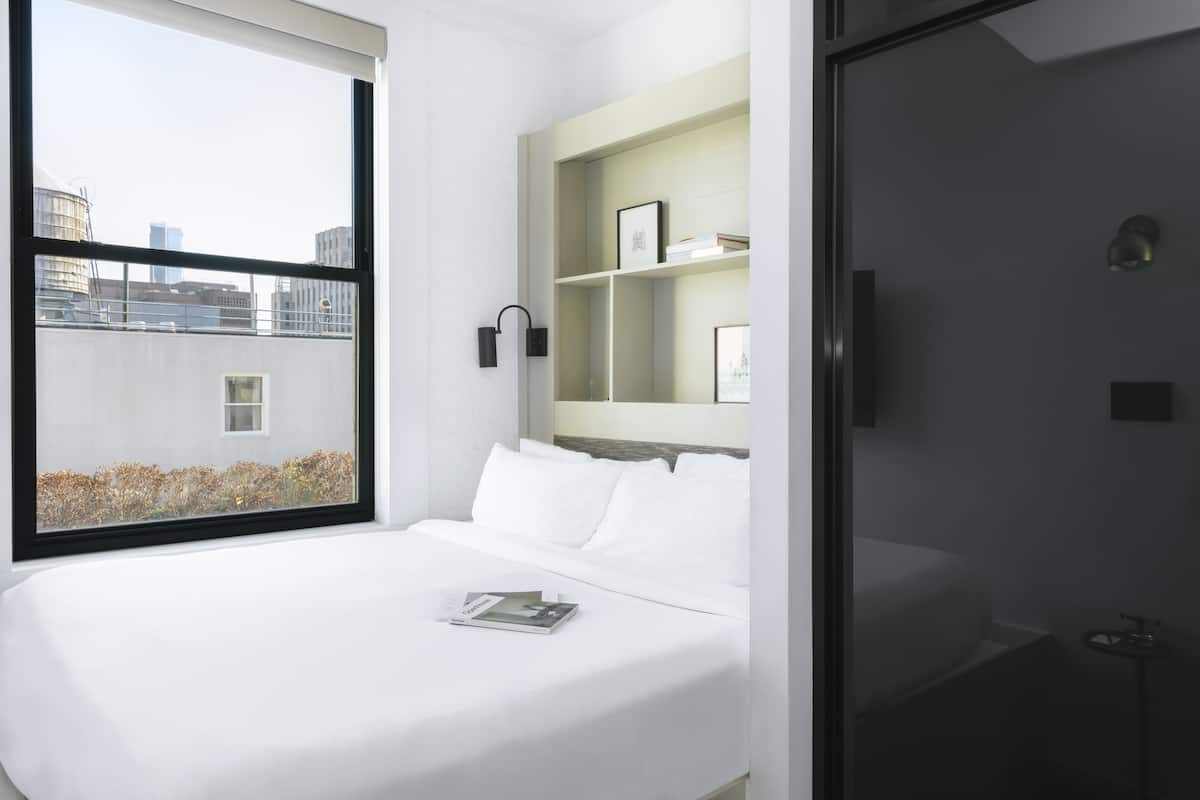 A modern hotel room with a large bed, a bedside table, a small bedside lamp, and a view of the city through a large window.