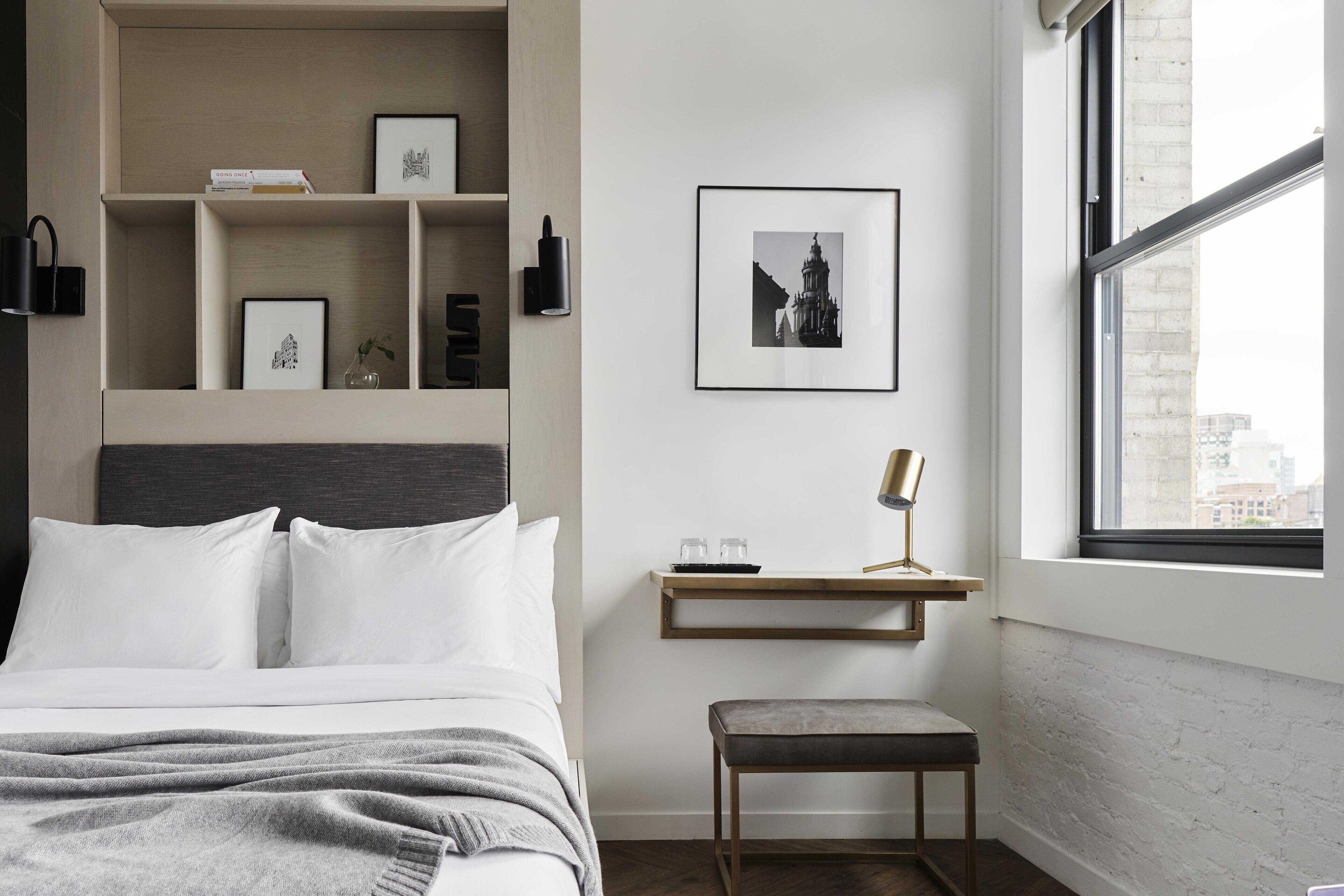 A bedroom with a bed, bedside table, and a window with a view of buildings.