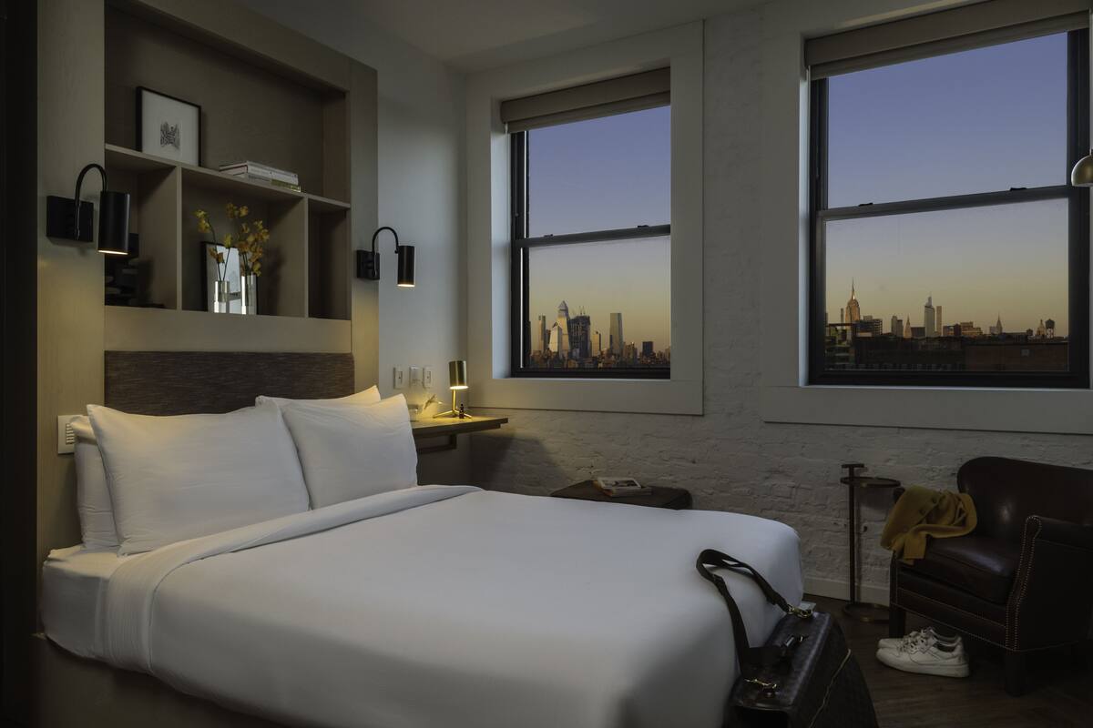 A hotel room with a large bed, a nightstand, a chair, and a view of a city skyline through two large windows.