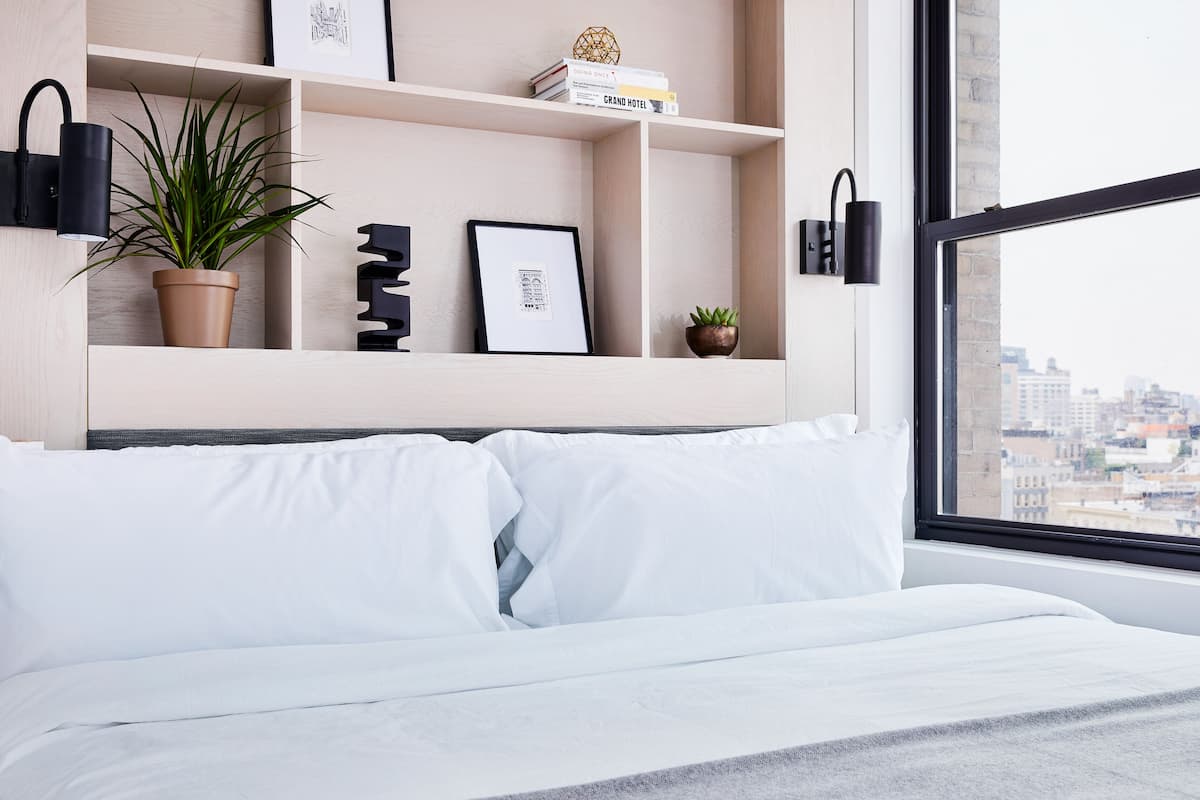 A modern hotel room with a bed, a city view, a bookshelf with books and decorative items, and wall-mounted lighting.