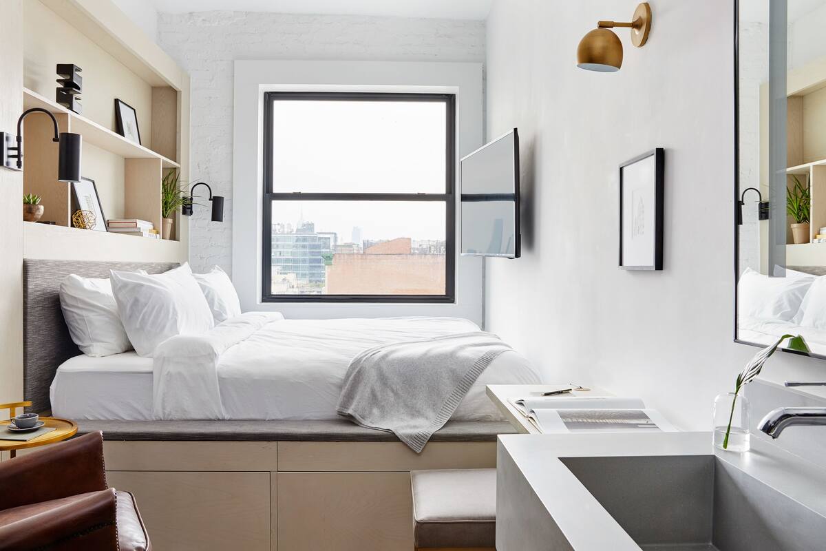 A modern hotel room with a large window, a bed with white linens, a built-in desk with a sink, and a city view.
