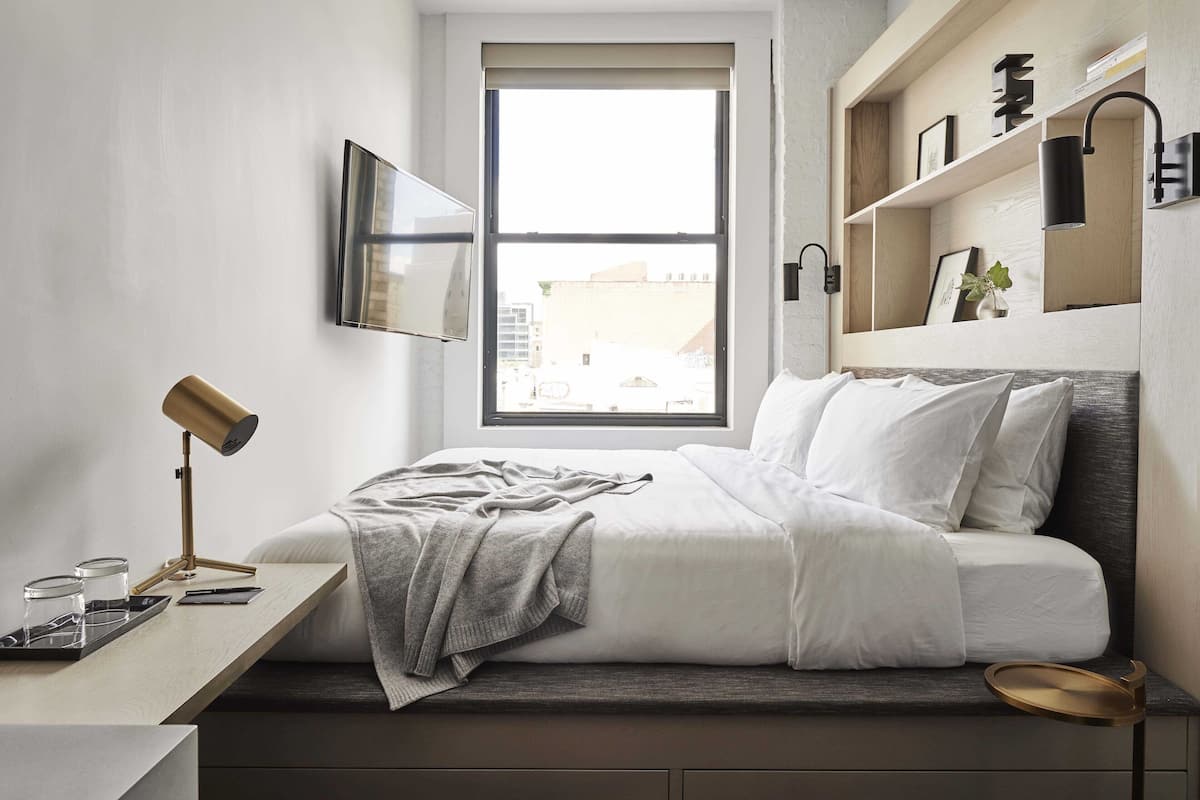 A modern bedroom with a bed, bedside table, lamp, and a window with a view of buildings.
