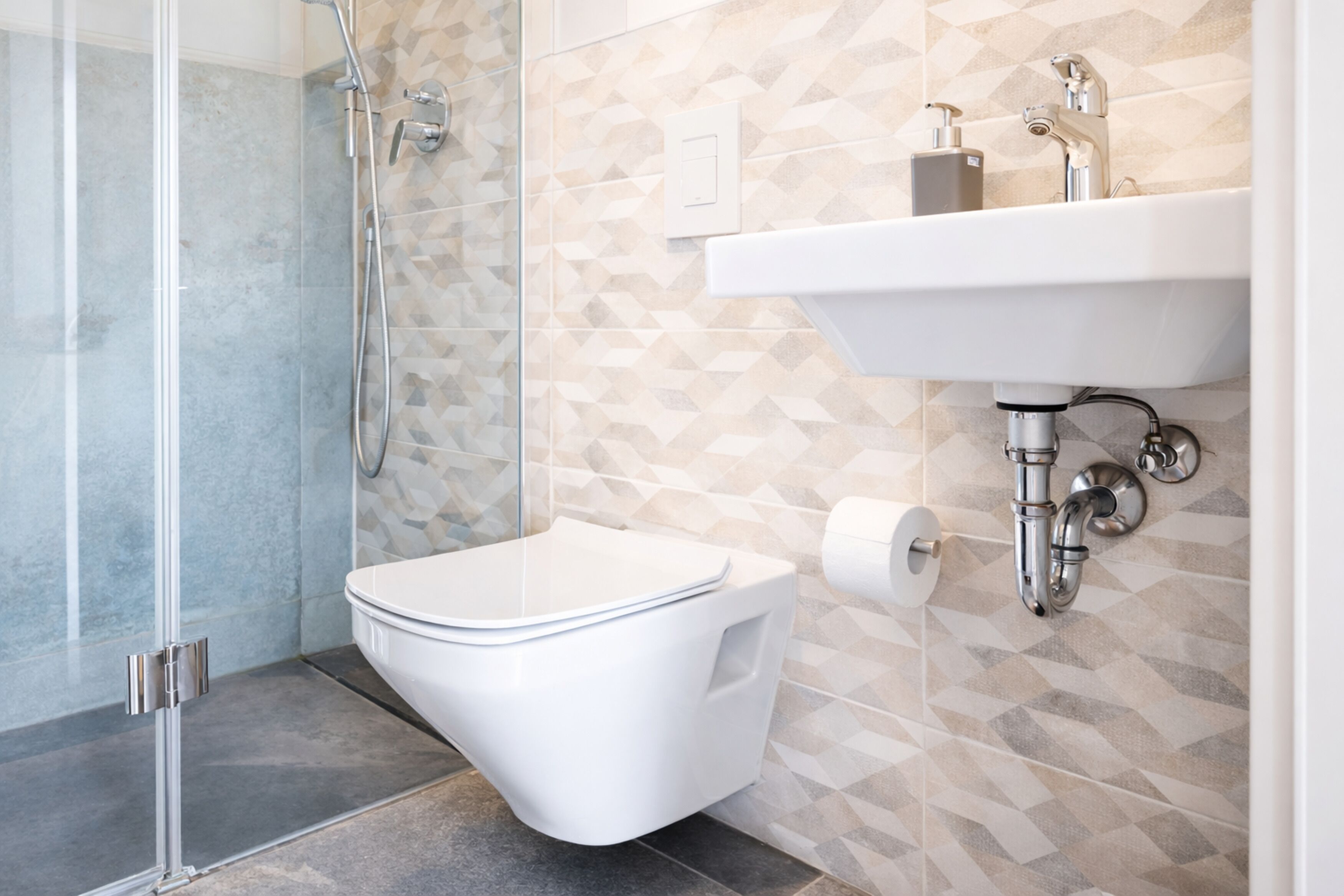 Un ba ño moderno con cabina de ducha de vidrio, inodoro blanco y lavamanos con salpicadero de azulejos geométricos.