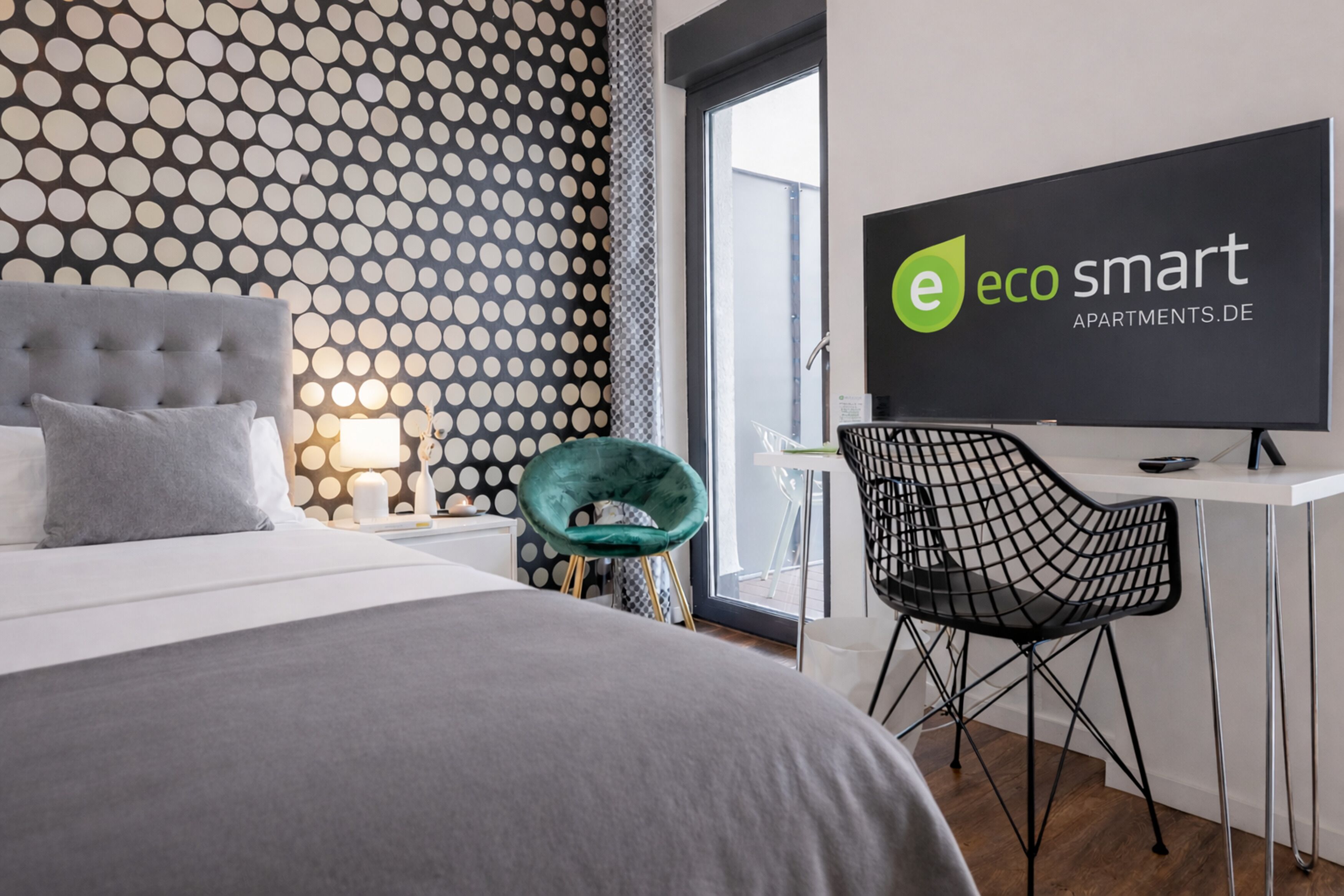 A modern hotel room with a polka dot wallpaper, a tufted headboard bed, a teal chair, a black wireframe chair, and a TV displaying 'eco smart apartments.de'.