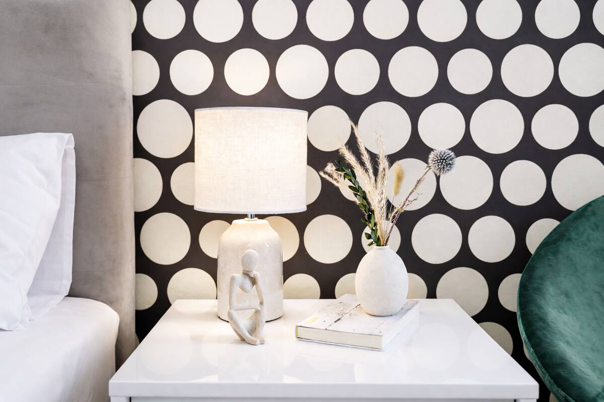 A neatly arranged bedroom with a white nightstand, a white lamp, a decorative vase with dried plants, and a polka dot wallpaper.