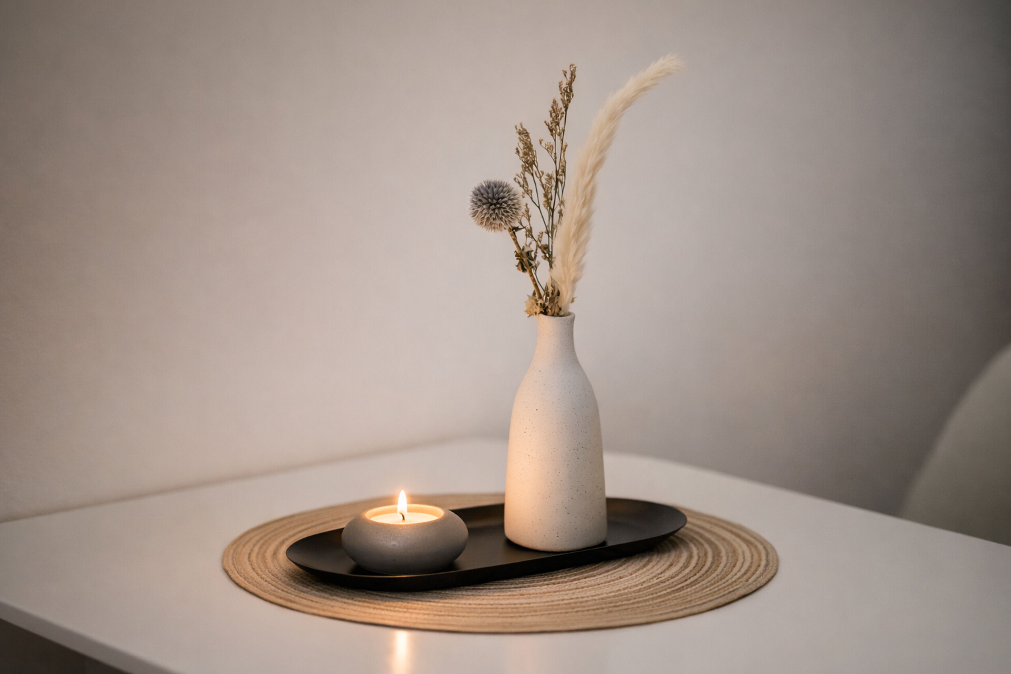 Una candela in un vaso bianco su un vassoio nero con un sottotavolo intrecciato, posizionata su una superficie bianca.