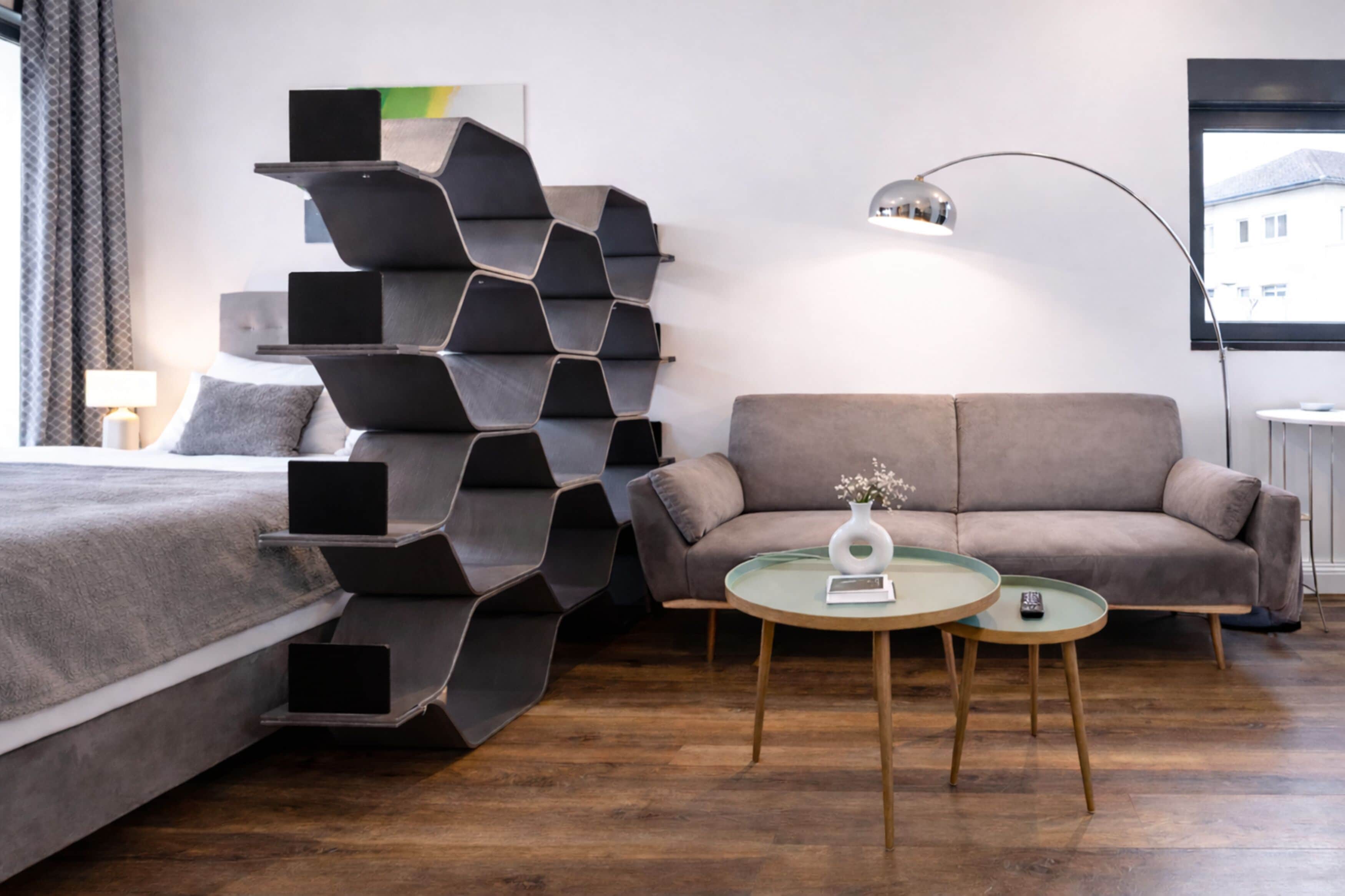 A modern hotel room with a bed, a sofa, a unique black shelving unit, a round table with a vase, and a floor lamp.