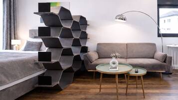 A modern hotel room with a bed, a sofa, a unique black shelving unit, a round table with a vase, and a floor lamp.