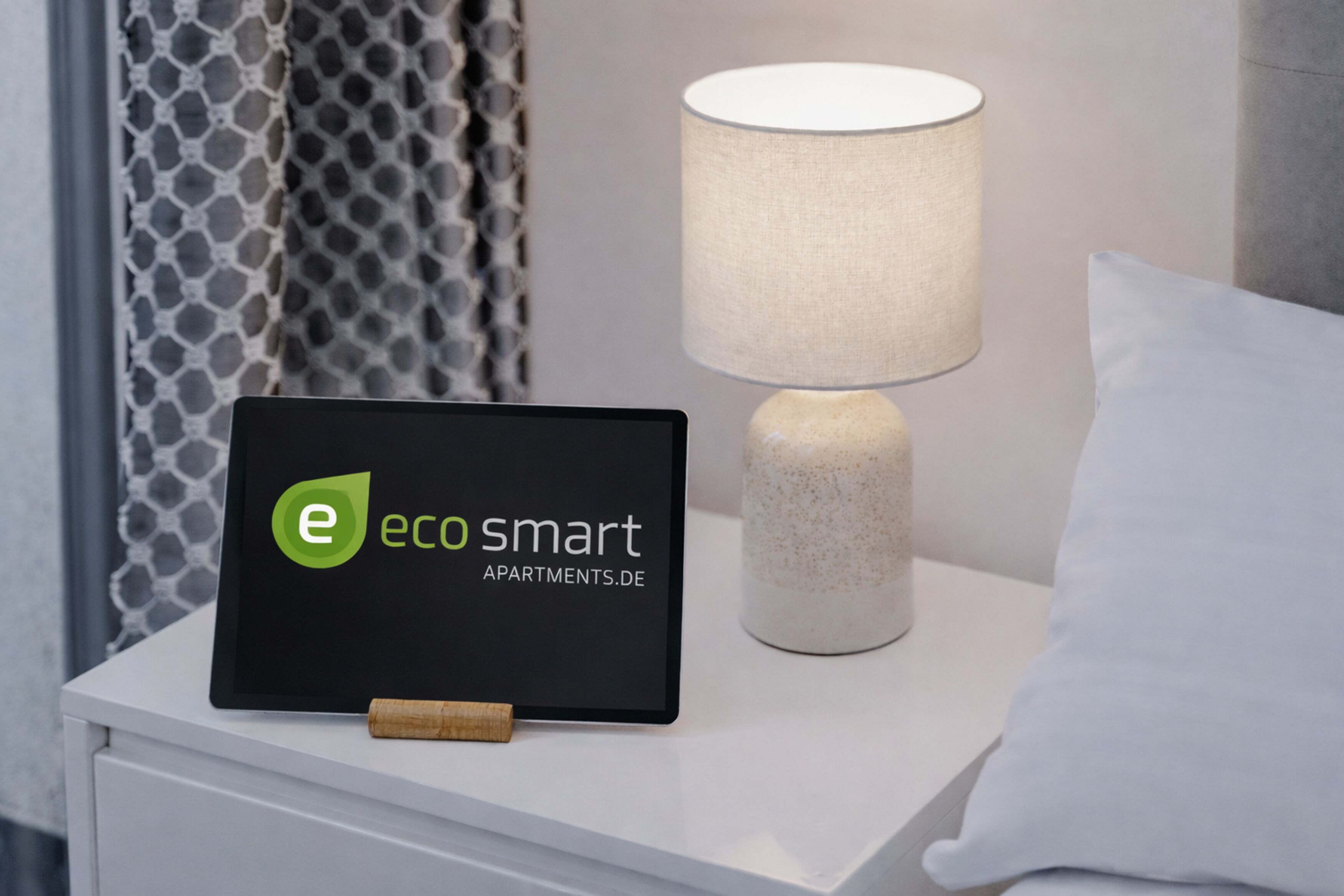 A tablet displaying 'eco smart apartments.de' on a bedside table with a lamp and a pillow.