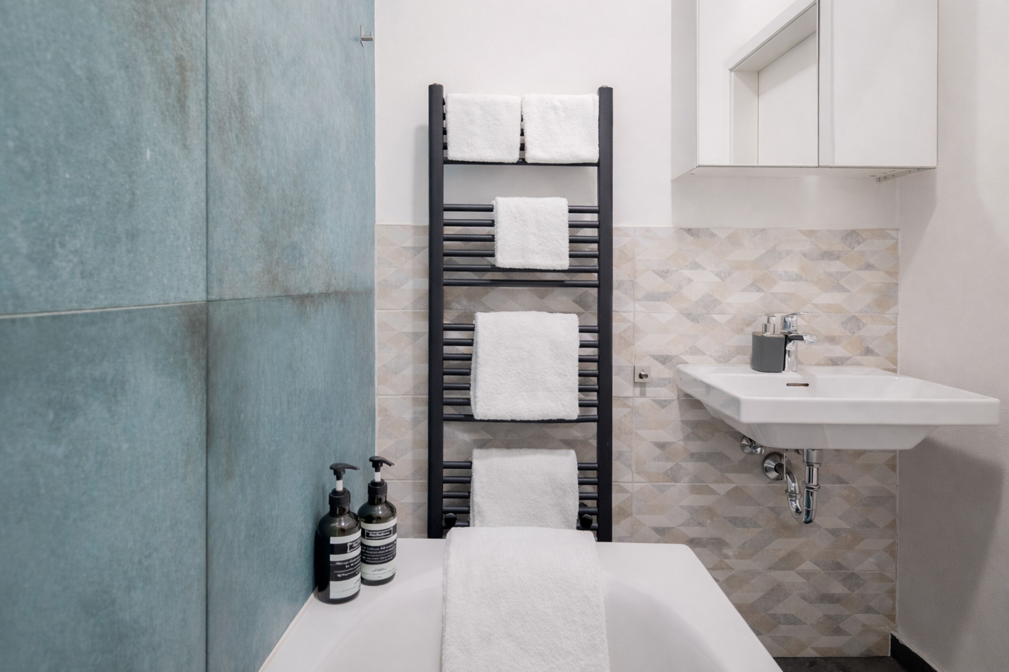 A modern bathroom with a black towel rack holding three towels, a white sink, and two bottles of hand soap.