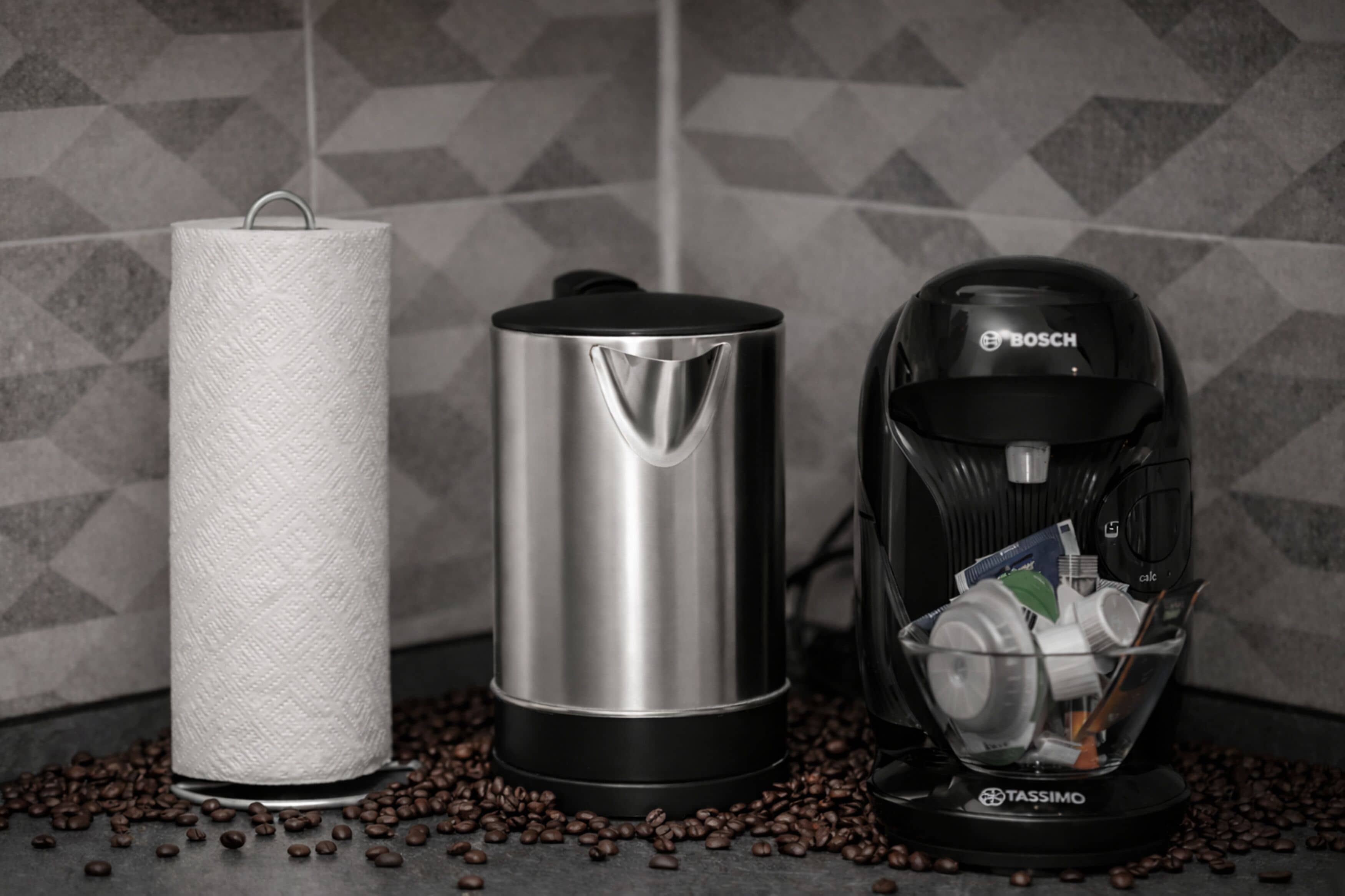 Une serviette en papier, un récipient en acier inoxydable et une machine à café dotée d’un compartiment pour les dosettes, le tout posé sur une surface où se trouvent des grains de café.