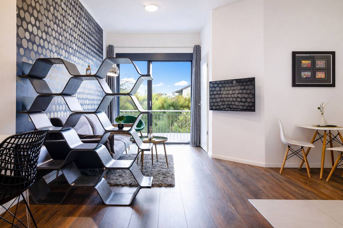A modern living room with a geometric wall design, a green chair, a small round table, and a TV on the wall.