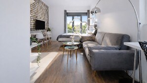 En moderne stue med en grå sofa, et rundt sofabord, et tv monteret på en geometrisk væg og et stort vindue med udsigt til grønne omgivelser.