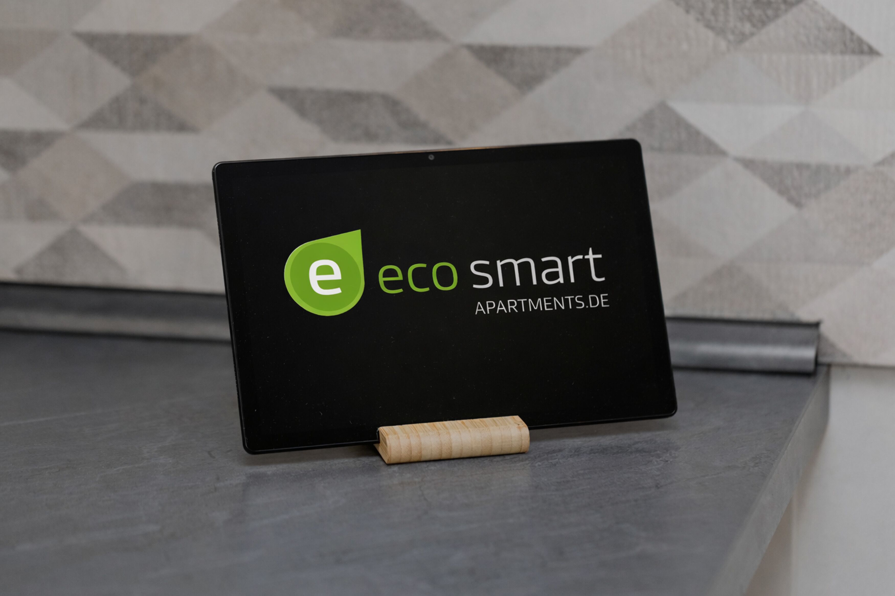 A tablet displaying the logo and name of 'eco smart apartments.de' on a black background with a geometric wall pattern in the background.