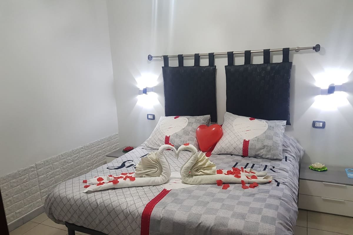 Een net opgemaakt bed met een geruit sprei, twee kussens en een kussen in hartvorm. Er zijn twee wandlampen en een klein tafeltje met een groen voorwerp erop.