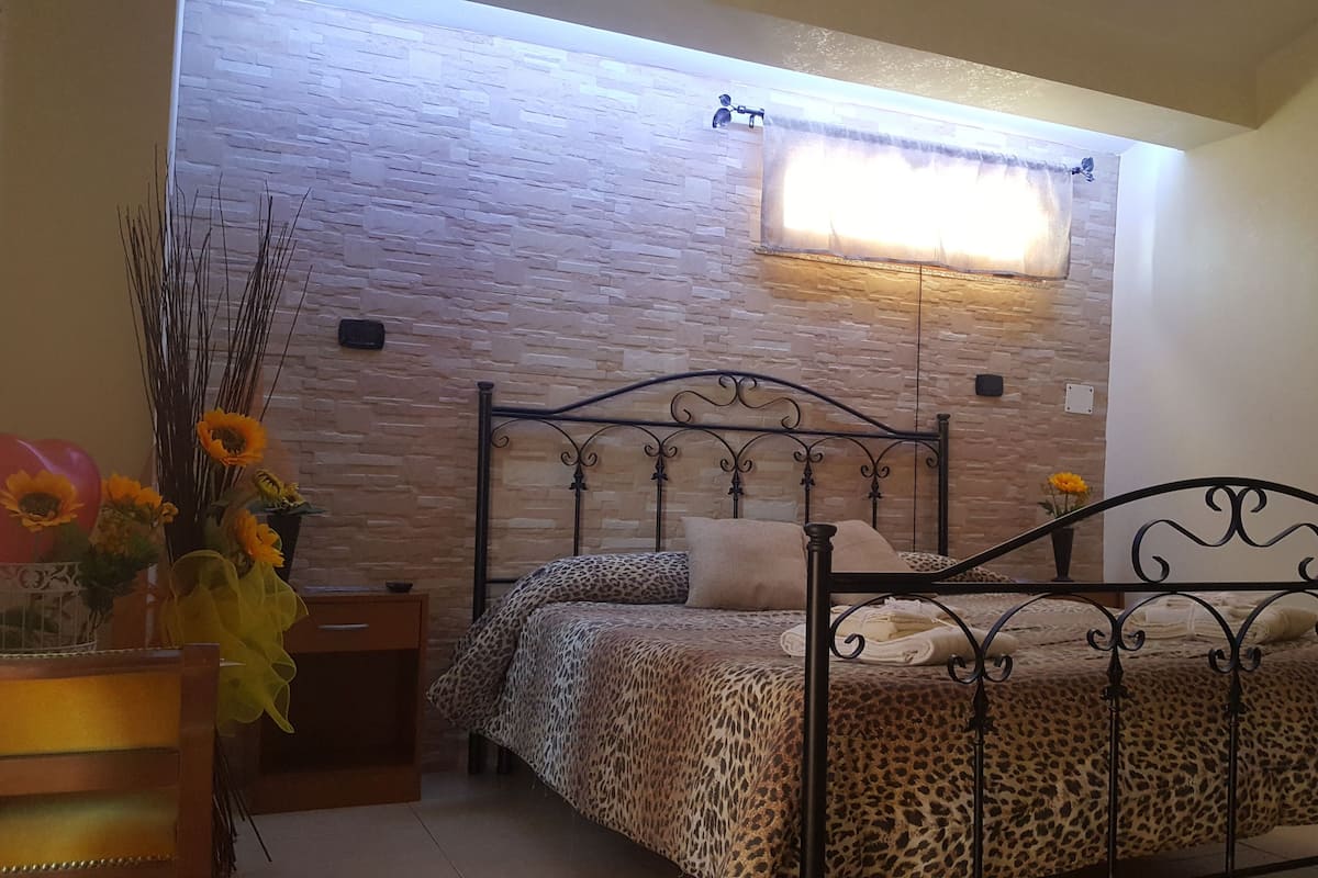 Een slaapkamer met een stenen muur, een bed met een luipaardprint hoes, een nachtkastje, een vaas met zonnebloemen en een raam met gordijn.
