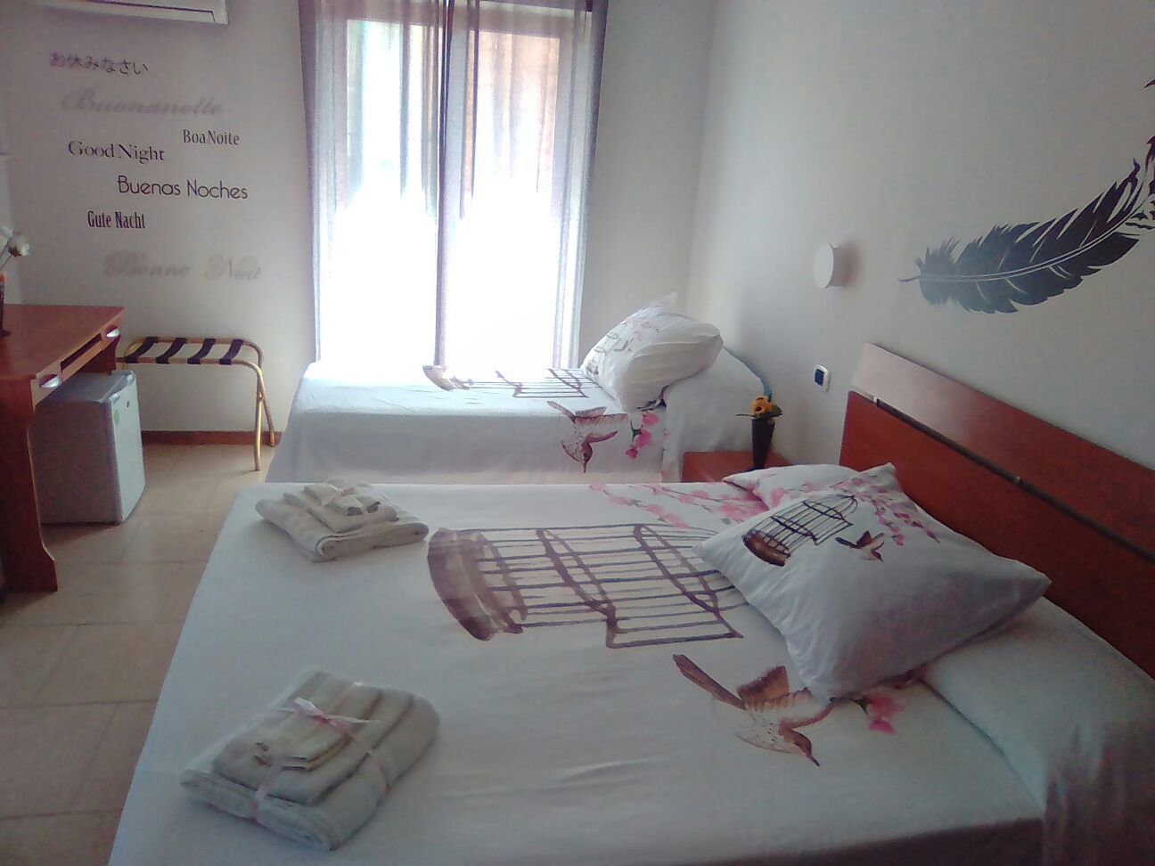 Une chambre d’hôtel avec deux lits, un bureau et une chaise. Il y a des serviettes sur les lits et une décoration en plumes accrochée au mur.