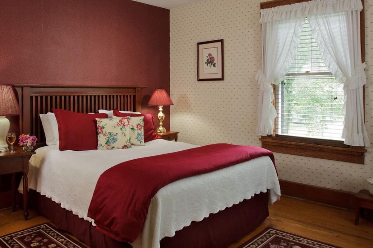 A bedroom with a wooden bed, red bedding, a nightstand with a lamp, a framed picture on the wall, and a window with curtains.