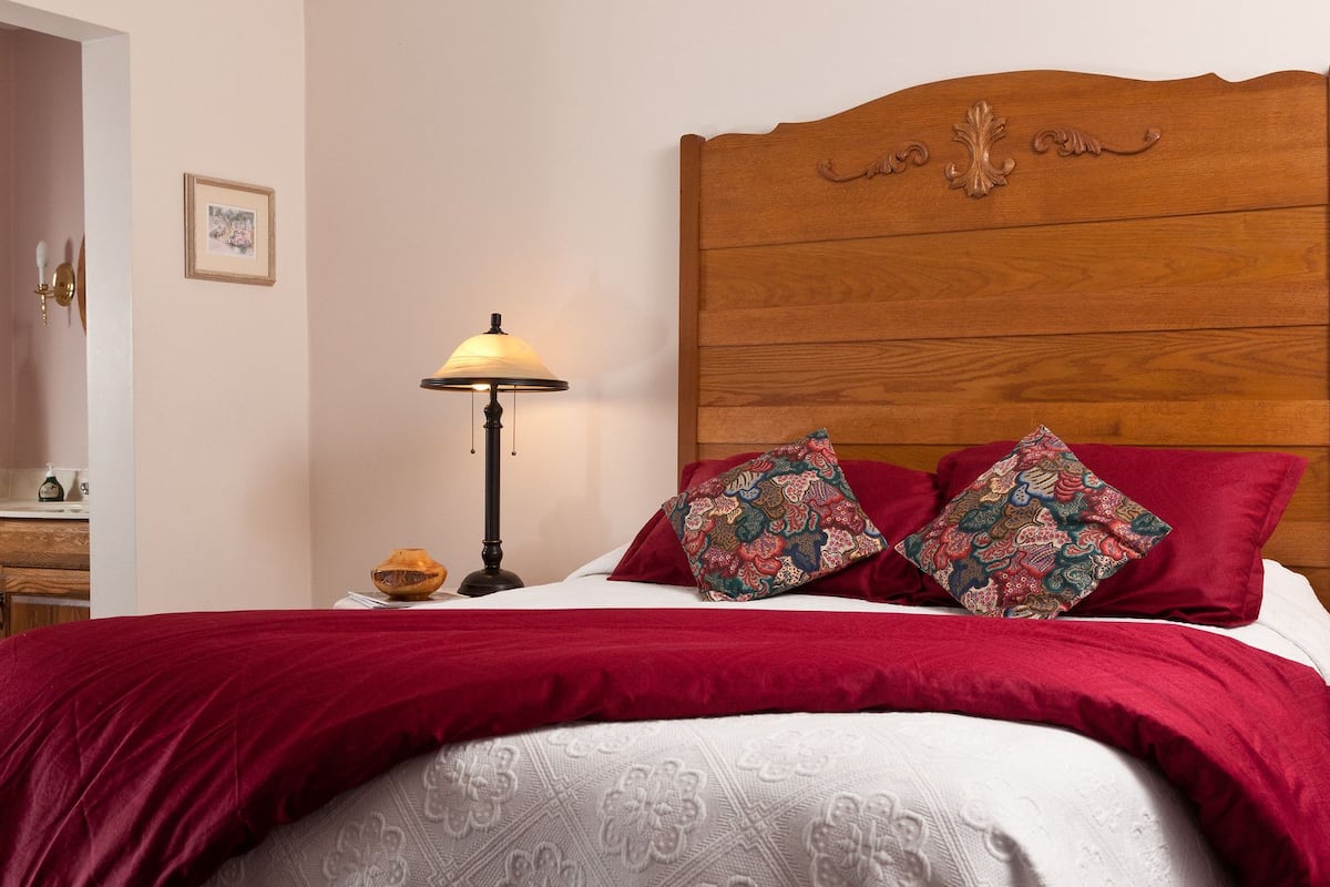 A neatly made bed with a red bedspread and two patterned pillows, a wooden headboard, a bedside lamp, and a small decorative bowl on the nightstand.