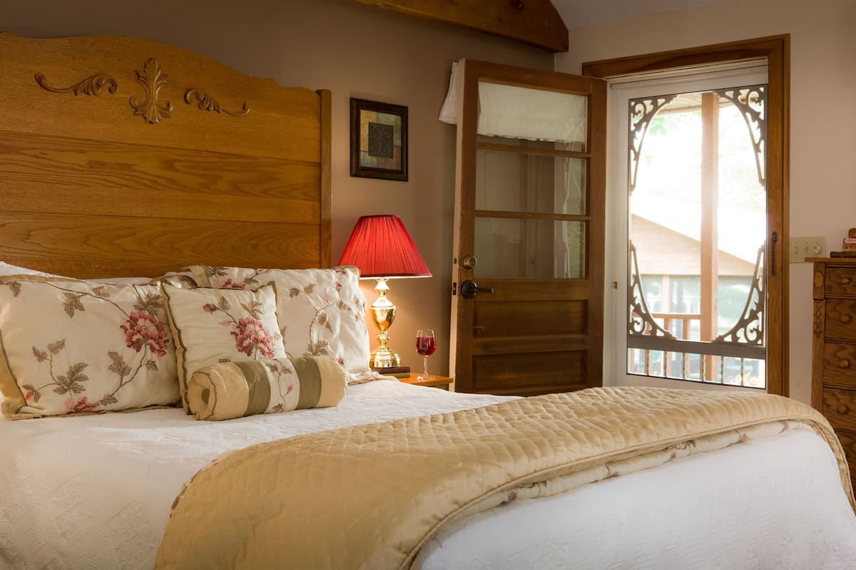 A bedroom with a wooden headboard, floral bedding, a red lamp, and a wooden door with a view.