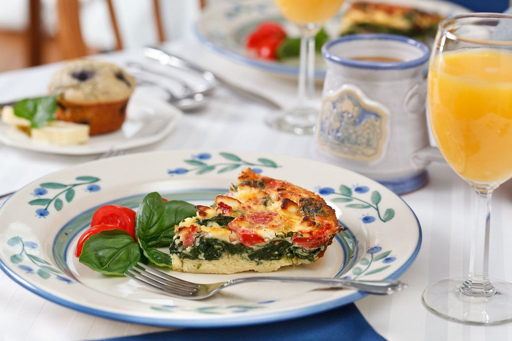 A breakfast setting with a slice of quiche on a floral plate, a muffin, a glass of orange juice, and a cup of coffee.