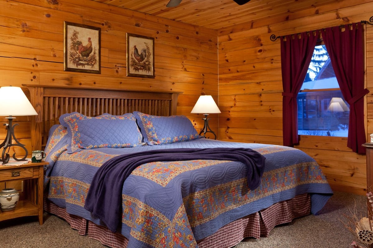 A cozy log cabin bedroom with a large bed, two bedside lamps, a wooden nightstand, and a window with curtains.