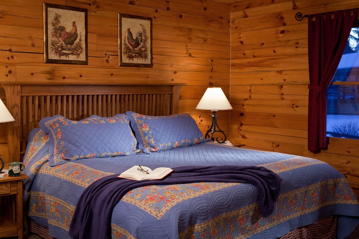 A wooden cabin room with a bed, two framed pictures on the wall, a nightstand, and a window with curtains.