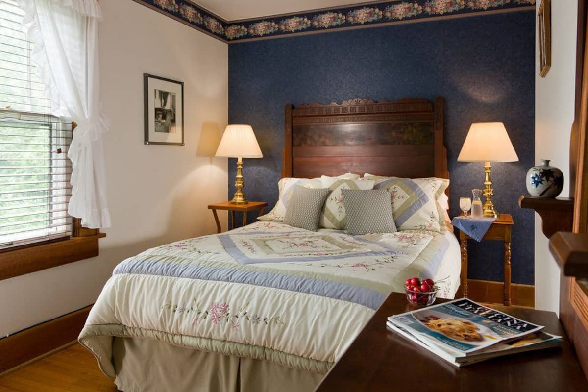 A bedroom with a bed, two bedside tables with lamps, a window with curtains, a framed picture on the wall, and a magazine on the bed.