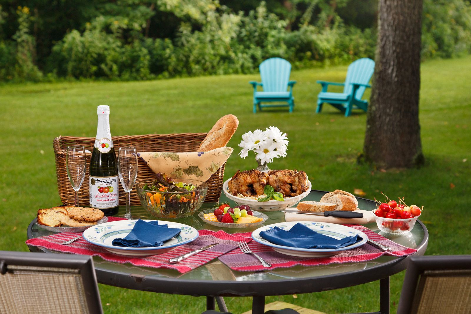 An outdoor dining setup with a table set for a meal, including a bottle of sparkling cider, a salad, grilled chicken, and various fruits and cheeses.