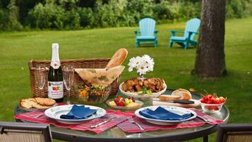 An outdoor dining setup with a table set for a meal, including a bottle of sparkling cider, a salad, grilled chicken, and various fruits and cheeses.