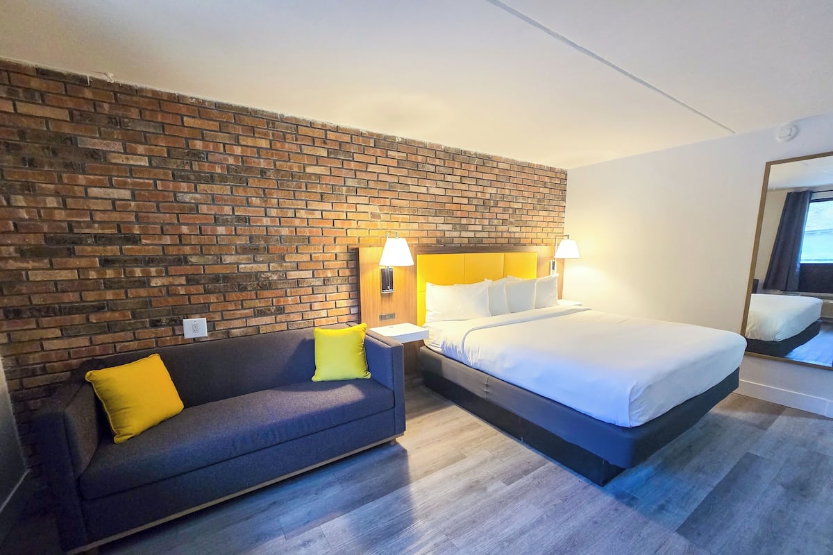 Quarto de hotel com parede de tijolos, cama com roupa de cama branca, sofá escuro com almofadas amarelas e mesa de cabeceira com abajur.