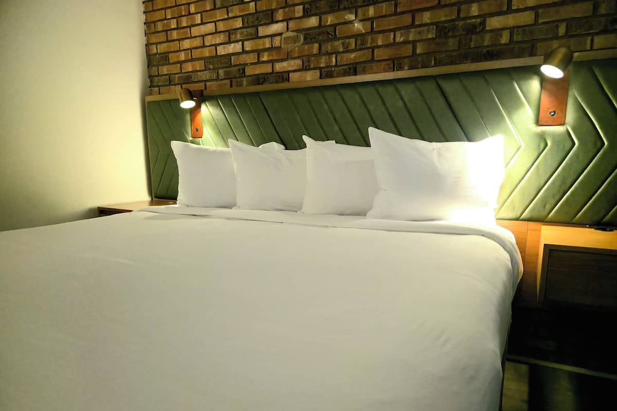 Cama bem arrumada com lençóis e almofadas brancos, cabeceira estofada verde e duas luminárias fixadas na parede.