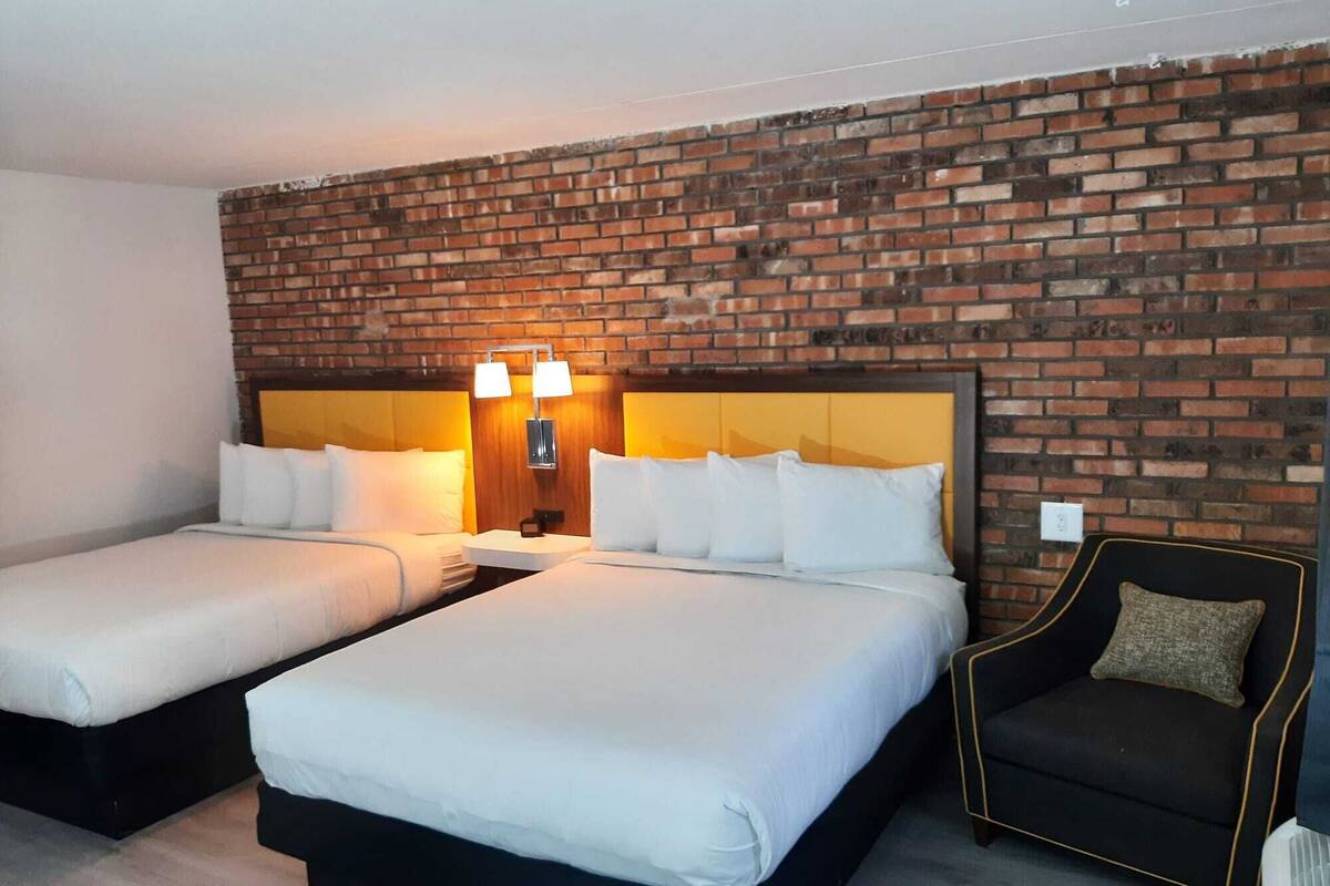 Quarto de hotel com duas camas, uma parede de tijolos e uma poltrona pequena.