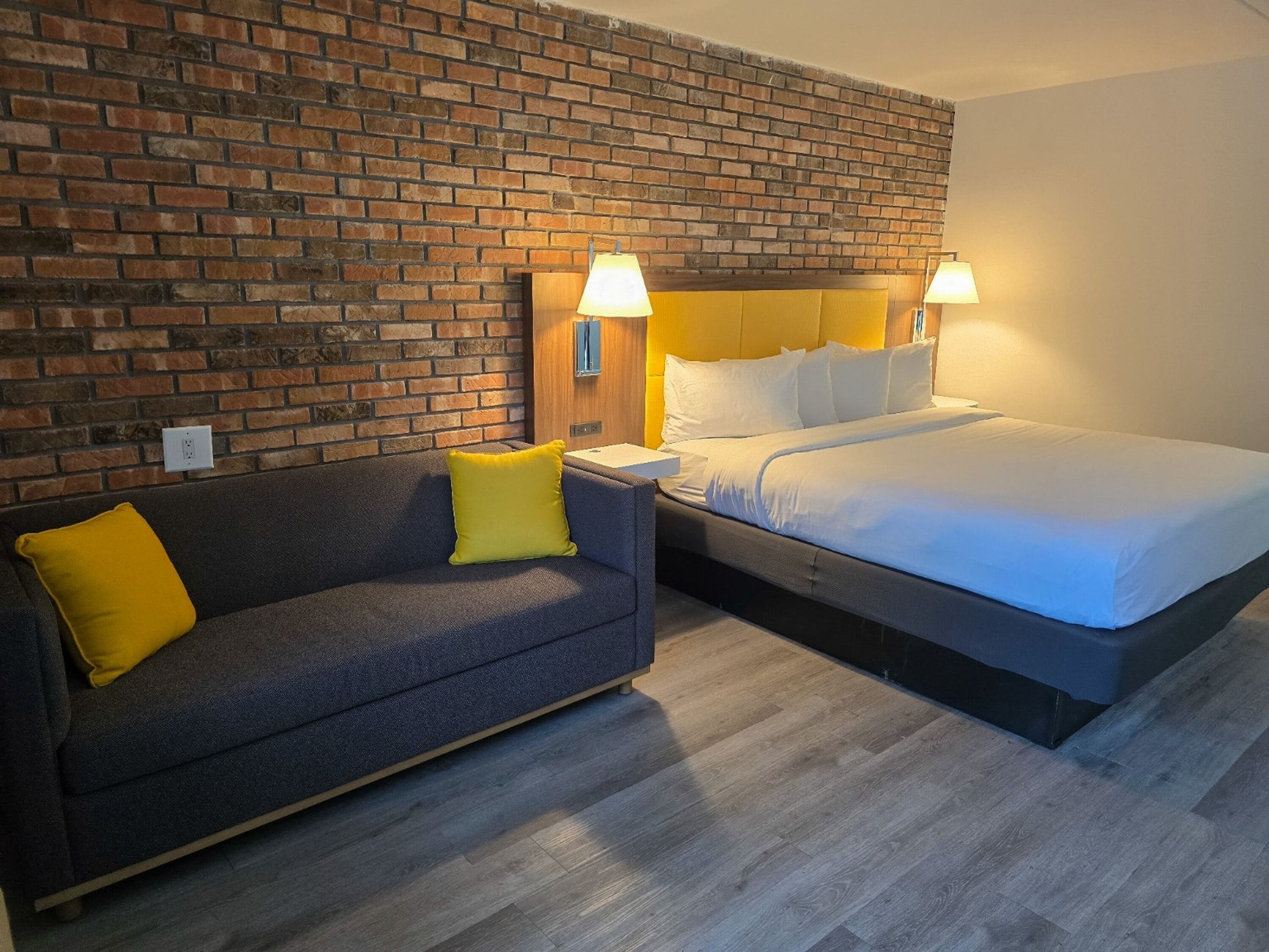 Una camera d'albergo con un muro in mattoni, un letto con biancheria bianca, un divano grigio con un cuscino giallo e due lampade da comodino.