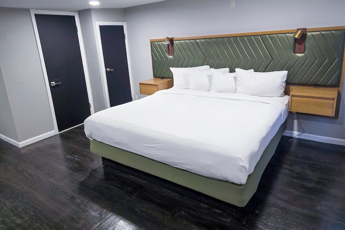 Quarto de hotel moderno com uma cama grande, mesa de cabeceira de madeira, painel de cabeceira estofado em verde e piso de madeira escura.