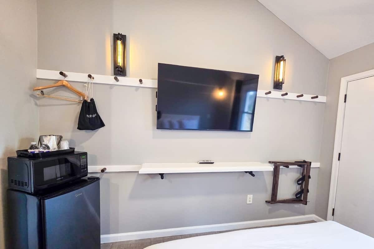 Quarto de hotel com televisão de tela plana na parede, micro-ondas, mini-geladeira, cama e prateleira de madeira com uma bolsa pendurada.