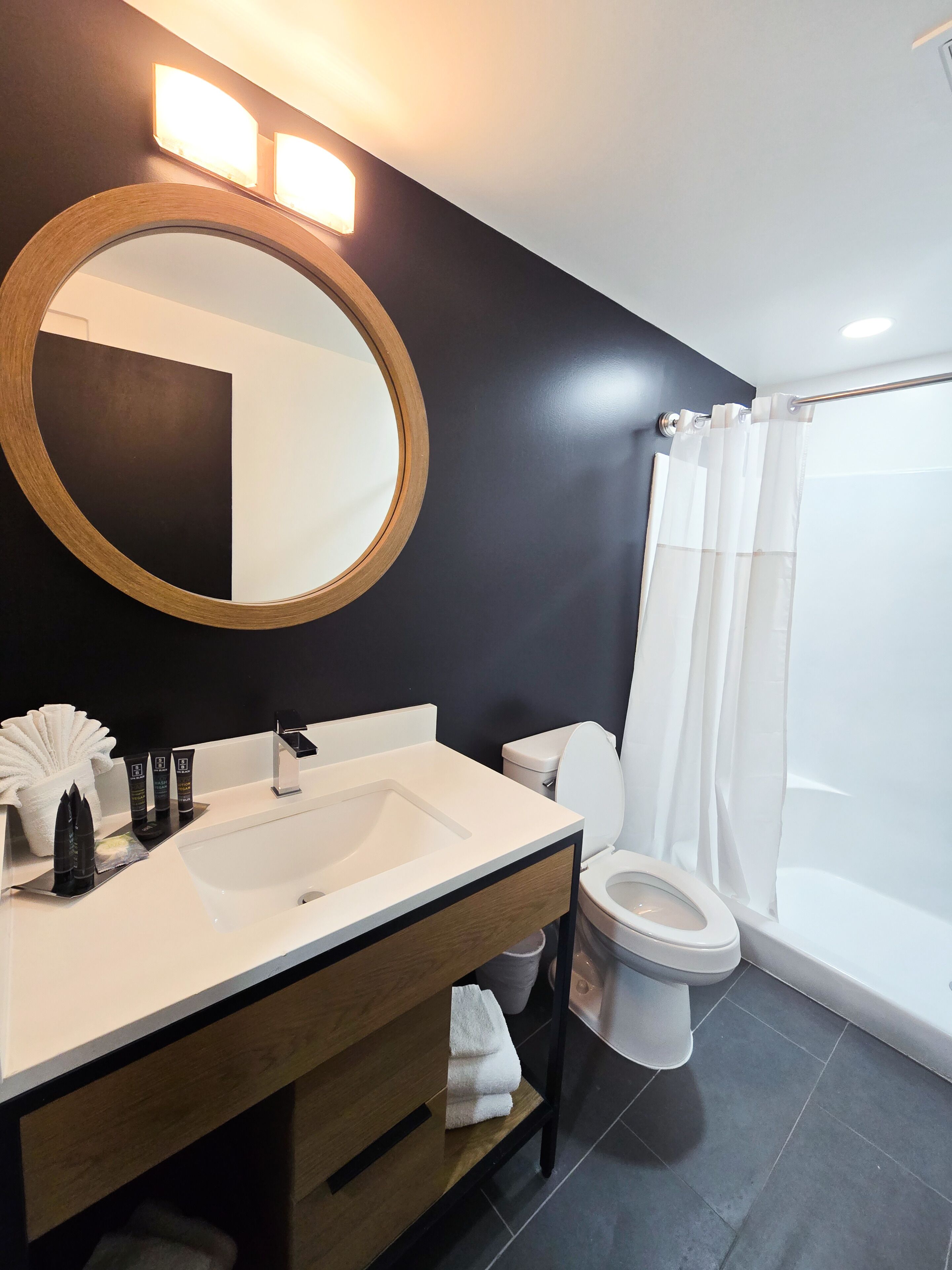 Une salle de bain moderne avec un grand miroir rond, un lavabo blanc, des toilettes et un espace douche.