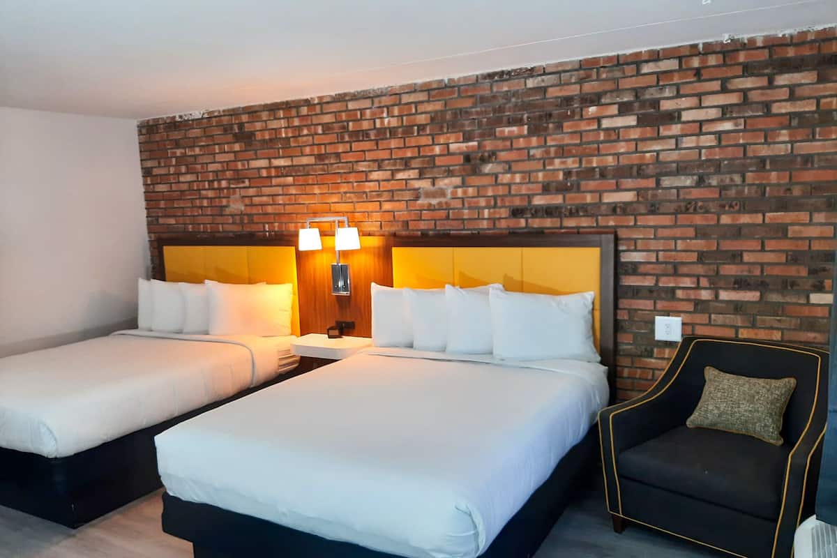 Quarto de hotel com duas camas, parede de tijolos, mesa de cabeceira com luminárias e uma poltrona preta.