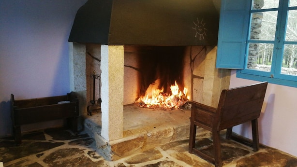 A stone fireplace with a fire burning inside, a wooden bench, and a chair in front of it. - O Albergue - Hostel (Lugo)