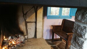 A stone fireplace with burning wood, a bench, and a window with blue shutters. - O Albergue - Hostel (Lugo)