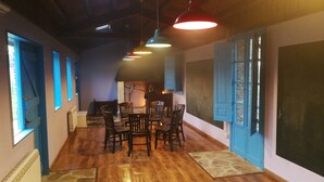 A dining area with wooden flooring, a round table, and chairs. There are multiple windows with blue shutters, and a blackboard on the wall. - O Albergue - Hostel (Lugo)