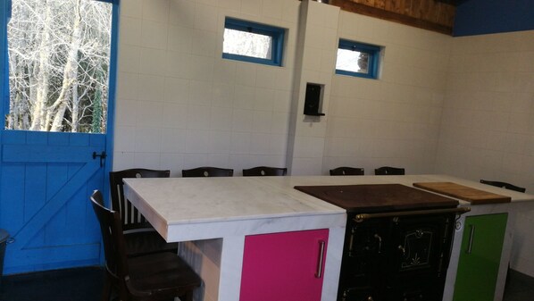 Una cocina con encimera blanca, un fregadero y armarios en tonos rosa, verde y negro. Hay dos ventanas con marcos azules y vistas a árboles.
