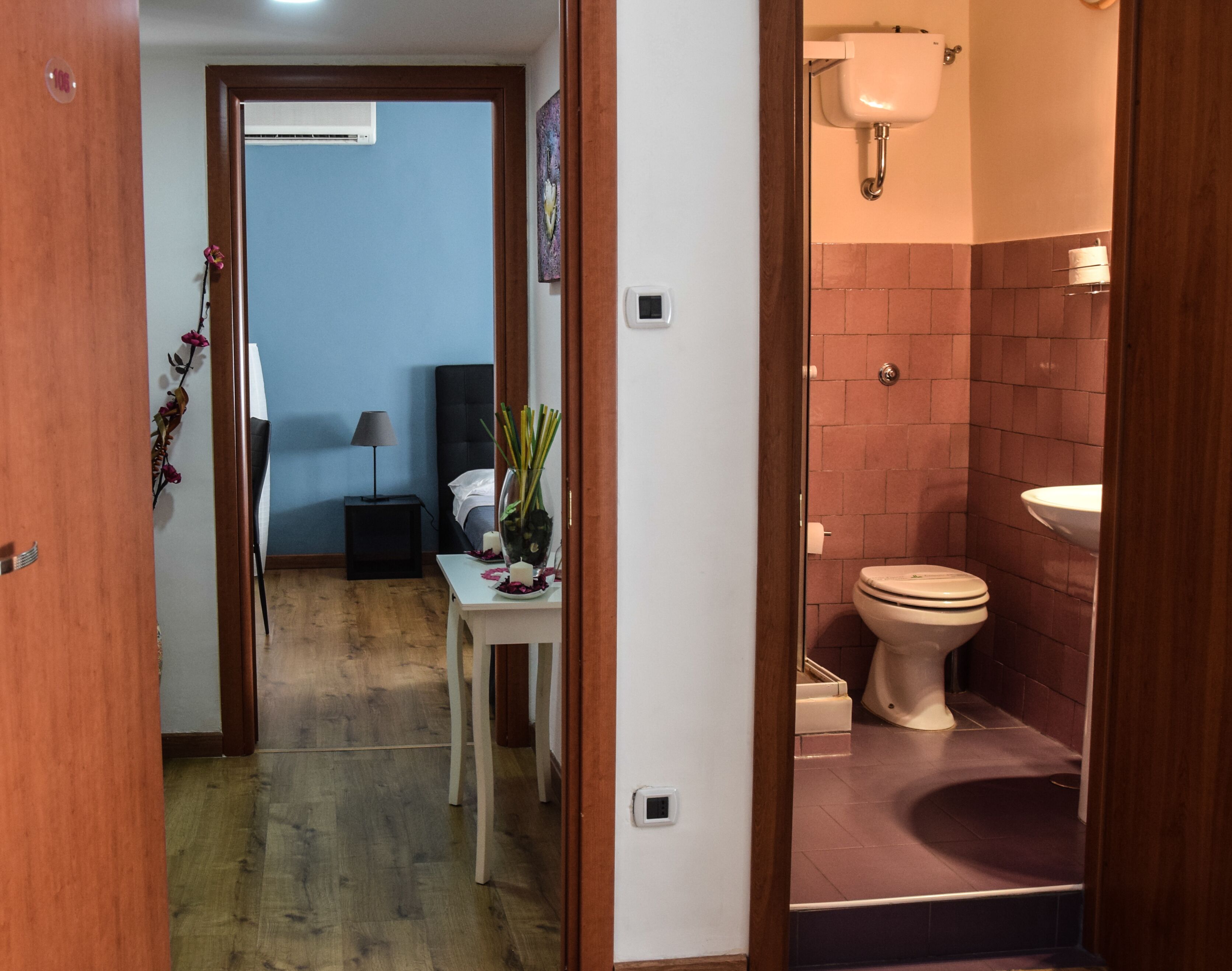 A hotel room with a wooden floor, a small table with a vase of flowers, a bed with a white headboard, and a bathroom with a toilet and sink.