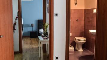 A hotel room with a wooden floor, a small table with a vase of flowers, a bed with a white headboard, and a bathroom with a toilet and sink.